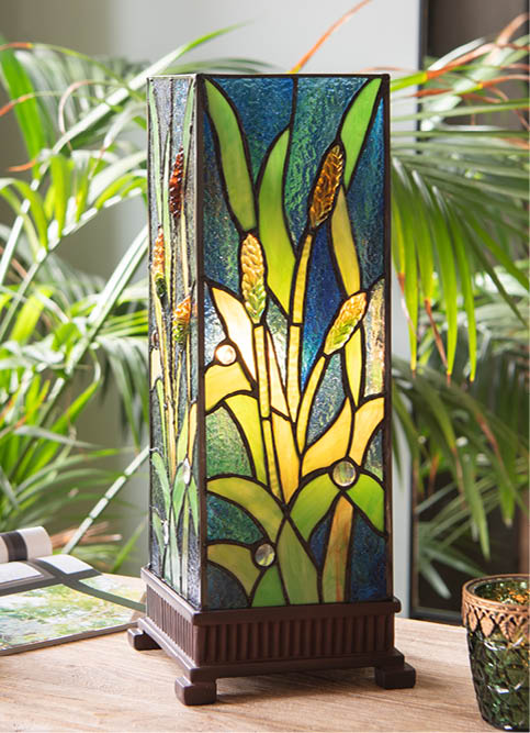 A table lamp with a stained glass design, standing on a wooden base. The glasswork features vibrant colors depicting an image of flowering plants and leaves, primarily in shades of green, yellow, and orange, providing a warm and natural appearance. The lamp is situated indoors in a room with lush green plants in the background, adding to the botanical atmosphere of the scene. The lamp's style lends an artistic and antique flair to the interior, and the lighting filtered through it would create a soothing glow in the space.