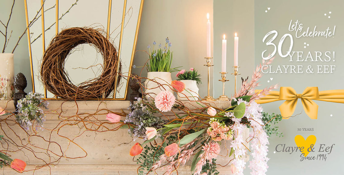 Een feestelijke decoratie die het 30-jarig jubileum viert van Clayre & Eef. Centraal in de compositie is een stijlvolle open haard met daarop een rijke schikking van bloemen en planten, waaronder roze tulpen en zachte bloesems, die samen een lenteachtige sfeer uitstralen. Een natuurlijke, krans van takken hangt tegen de spiegel, toevoegend aan het rustieke en huiselijke thema. Verspreid over de mantel staan elegante kaarsen in gouden kandelaars, die een warme gloed werpen op de omgeving. De scene is versierd met een groot, goudkleurig lint met een strik, en de woorden "Let's Celebrate! 30 Years! CLAYRE & EEF" zijn prominent in sierlijke witte letters aan de bovenkant van de afbeelding weergegeven, wat een uitnodigende en feestelijke sfeer creëert. Het algehele beeld is er een van elegantie en viering, passend voor een speciale mijlpaal.